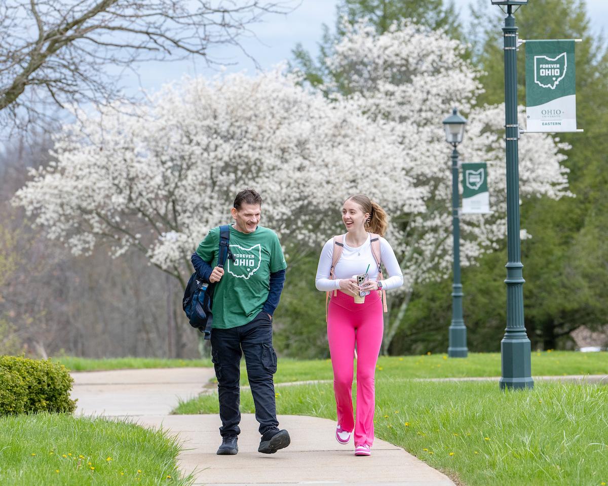 Two students walking down the sidewalk talking in front of blooming trees and OHIO flag.