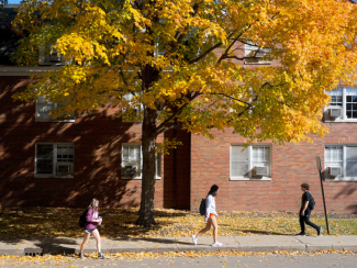 Students walking around campus in front of the brick buildings, sidewalks, and bright fall leaves. 