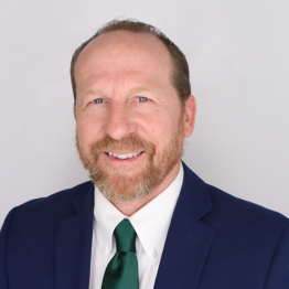 Curt Plummer in a blue suit with a white undershirt and green tie smiling in front of a white background.