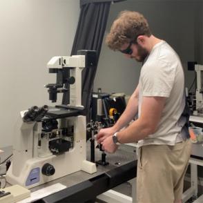 Hunter Thomas working on the Optical Tweezer setup, holding a small sheet that shows the infrared light of the laser when hit in front of a mirror to better align it. Once the laser is centered on the mirror, he will adjust the tilt of the mirror to reflect to the next one.