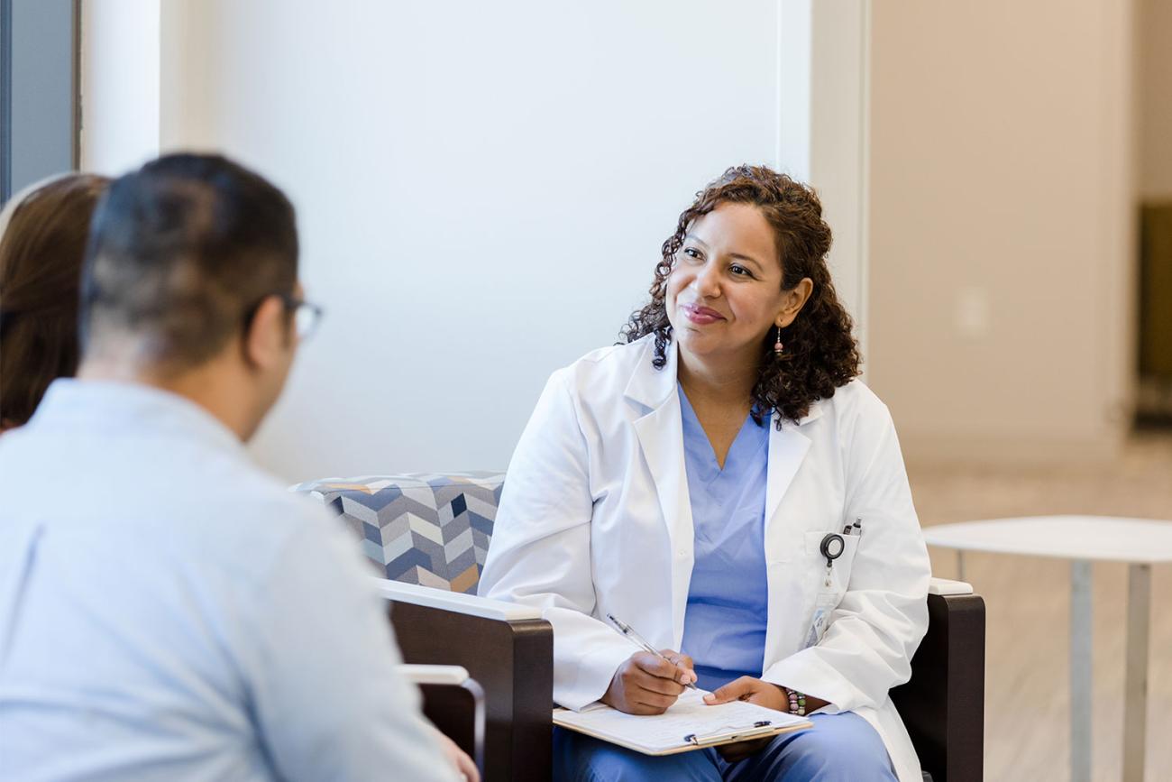 Woman with curly hair in white lab coat sitting with clipboard on her lap smiling at patient