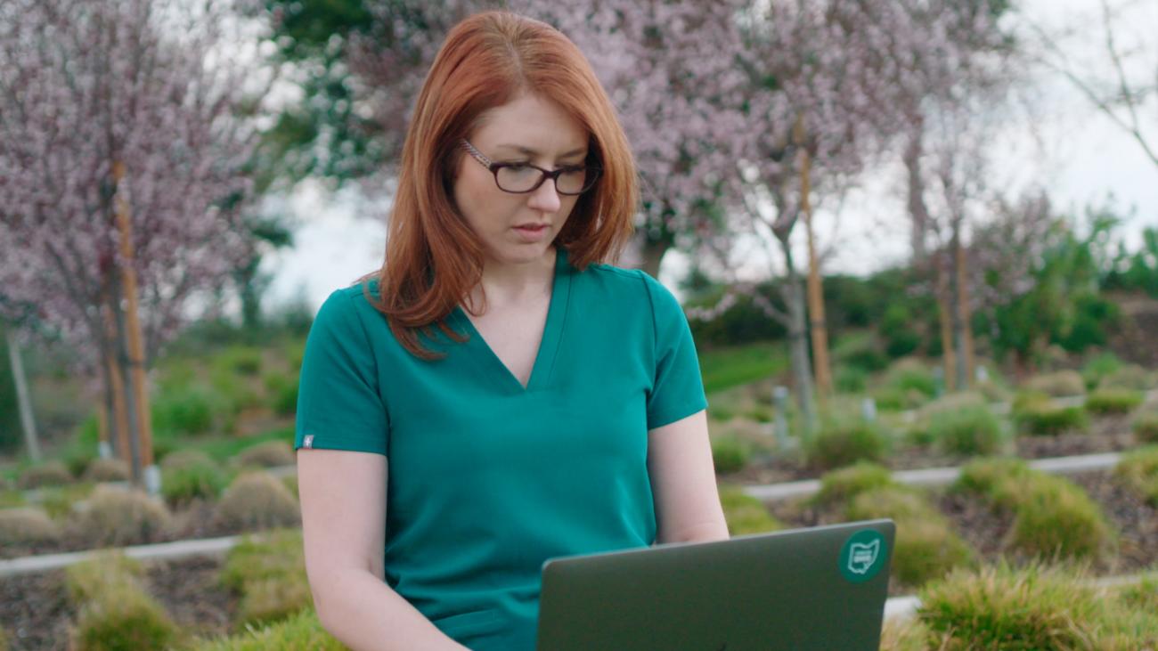 Woman in scrubs sitting outside looking at laptop