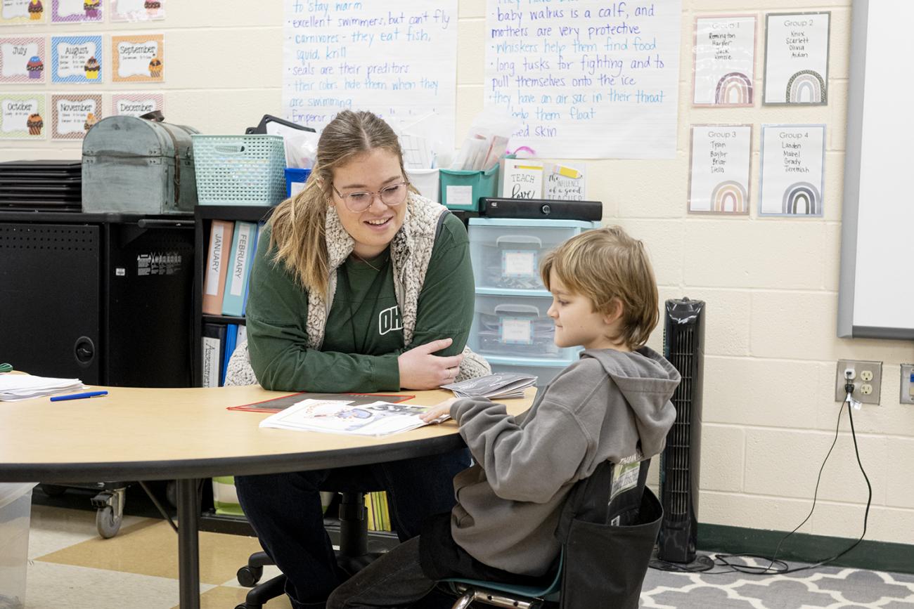 Teacher sits with child listening to him read a book.