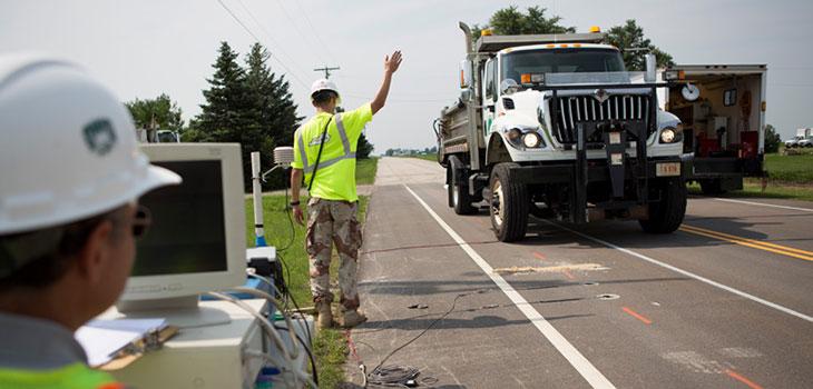 Road worker directs a dump truck on a rural two-lane highway while another worker stands next to a computer connected to cables extending onto the road