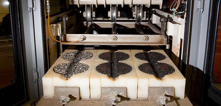 Rows of pieces of asphalt embedded in holes in the tops of smooth blocks, with a metal apparatus overhead aligned with the rows