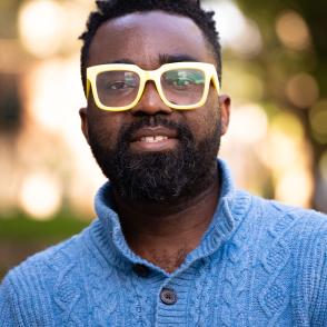 Raymond Ayivor looking at the camera and smiling, wearing a blue button-up sweater and yellow-framed glasses