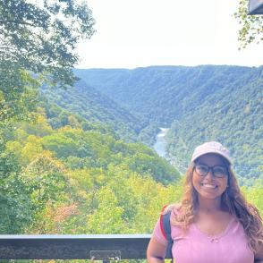 A photo of a woman in a pink shirt in front of a landscape.