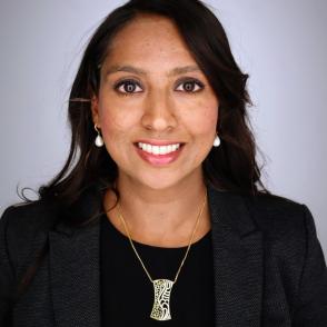 A photo of a professionally dressed woman, dressed in all black, in front of a gray background.