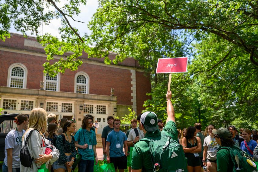 A group of individuals stands on college green, in front of the large brick memorial auditorium. One student stands in the middle of the group with a backpack, green polo, and a sign for the color group "red." 