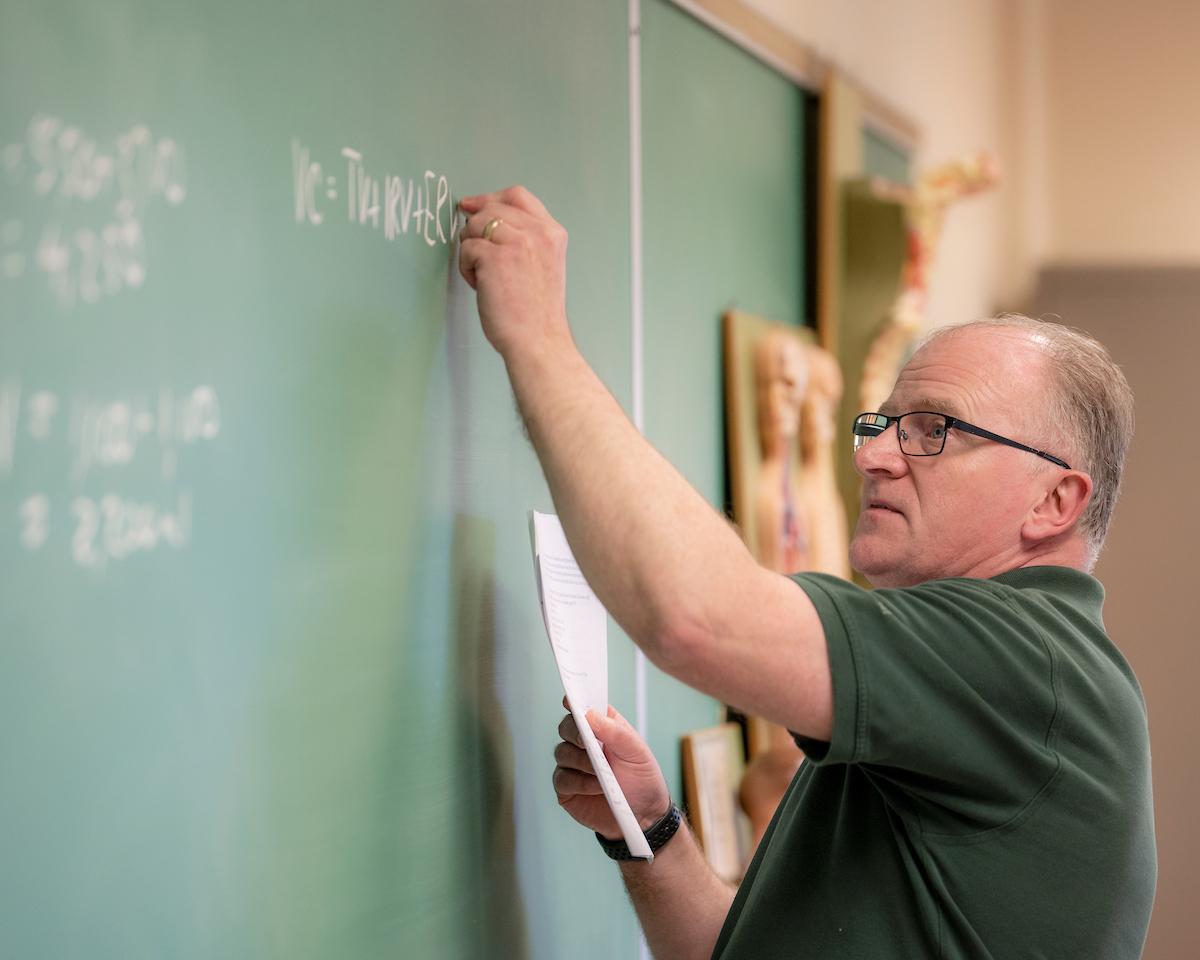Dr. Mark Waters writing an equation on a green chalkboard using white chalk.