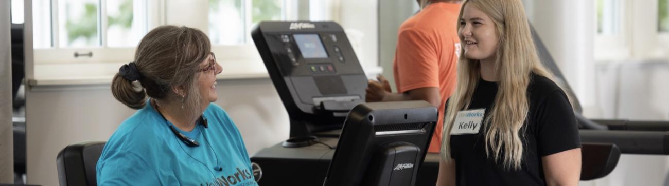 Student personal trainer at WellWorks speaking with a client seated on a cardio machine.