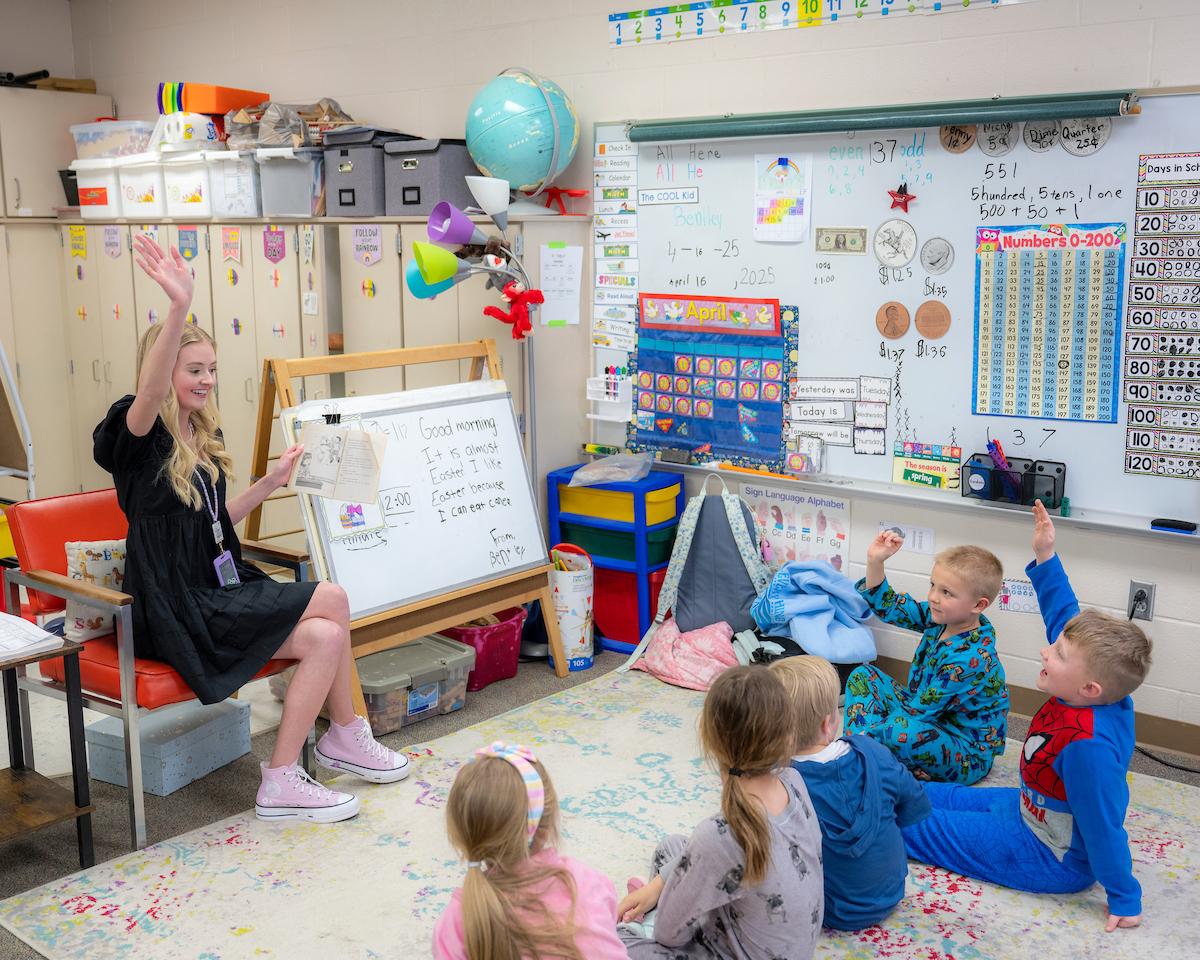 Female teacher, instructing children who are seated in a semi circle, sitting on a rug with their hands raised