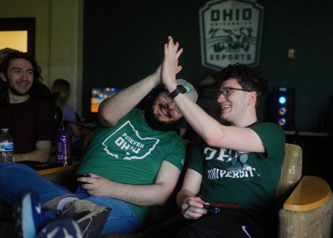 Two men wearing green Ohio University t-shirts holding video game remotes, high fiving each other