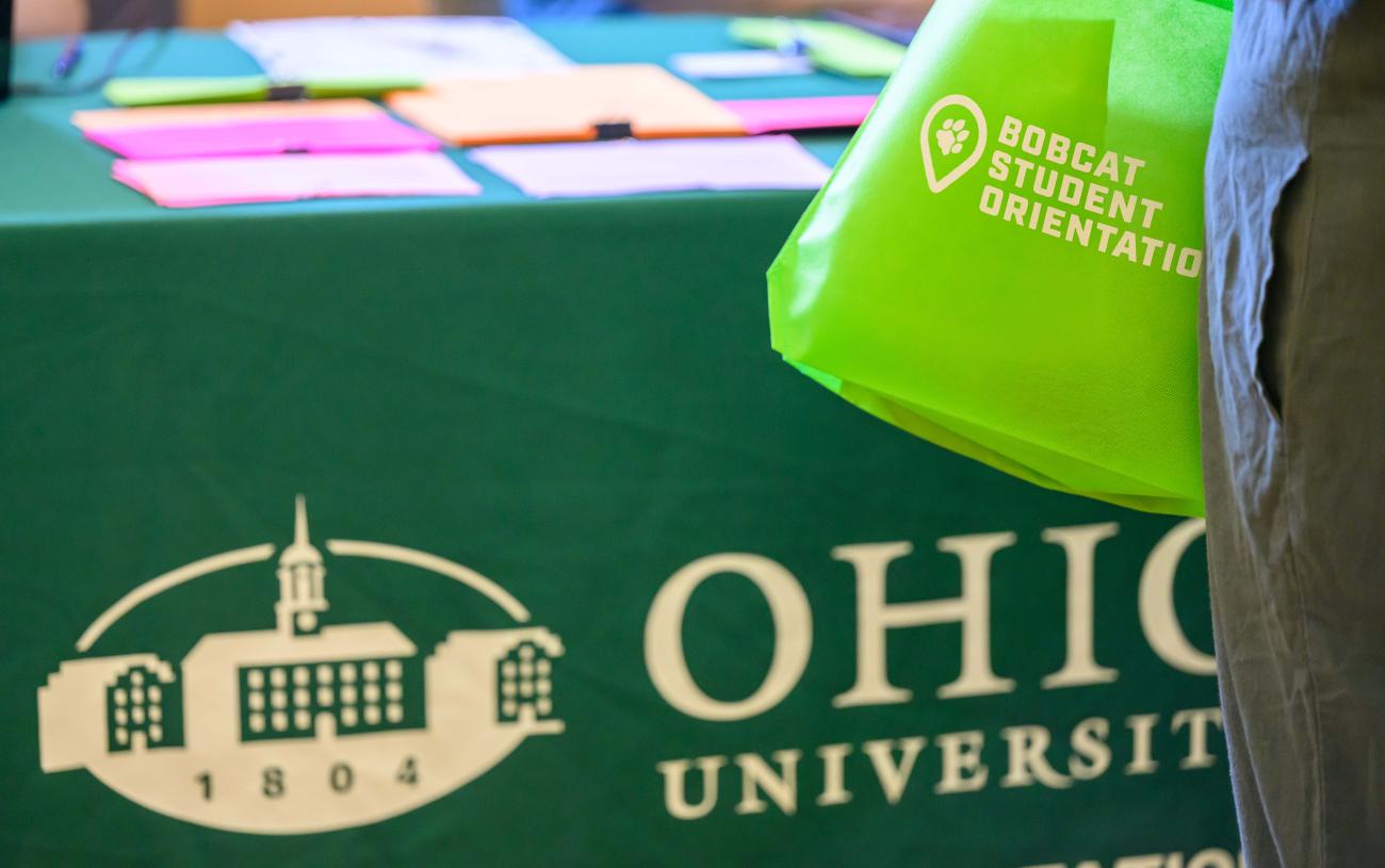 Someone holds a bright green bag that says Bobcat Student Orientation on it in front of a table covered by a darker green Ohio University table cloth with colorful paper sitting on it. 