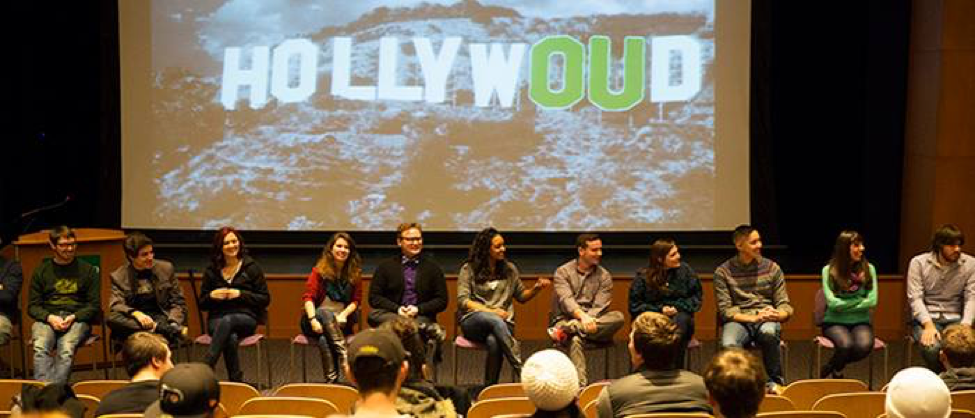 Panel of people sitting in chairs facing audience with screen behind that says "HollywOUd."