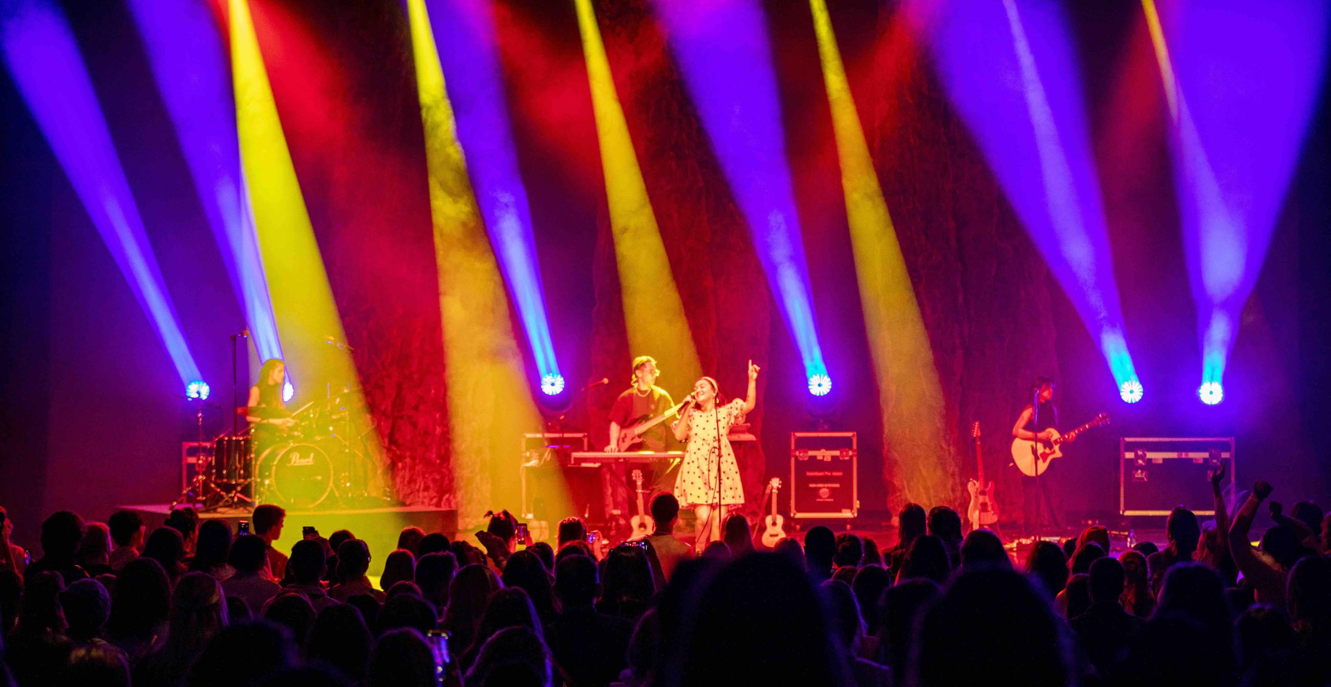 Crowd watching mxmtoon and three piece band perform with stage lights shining down