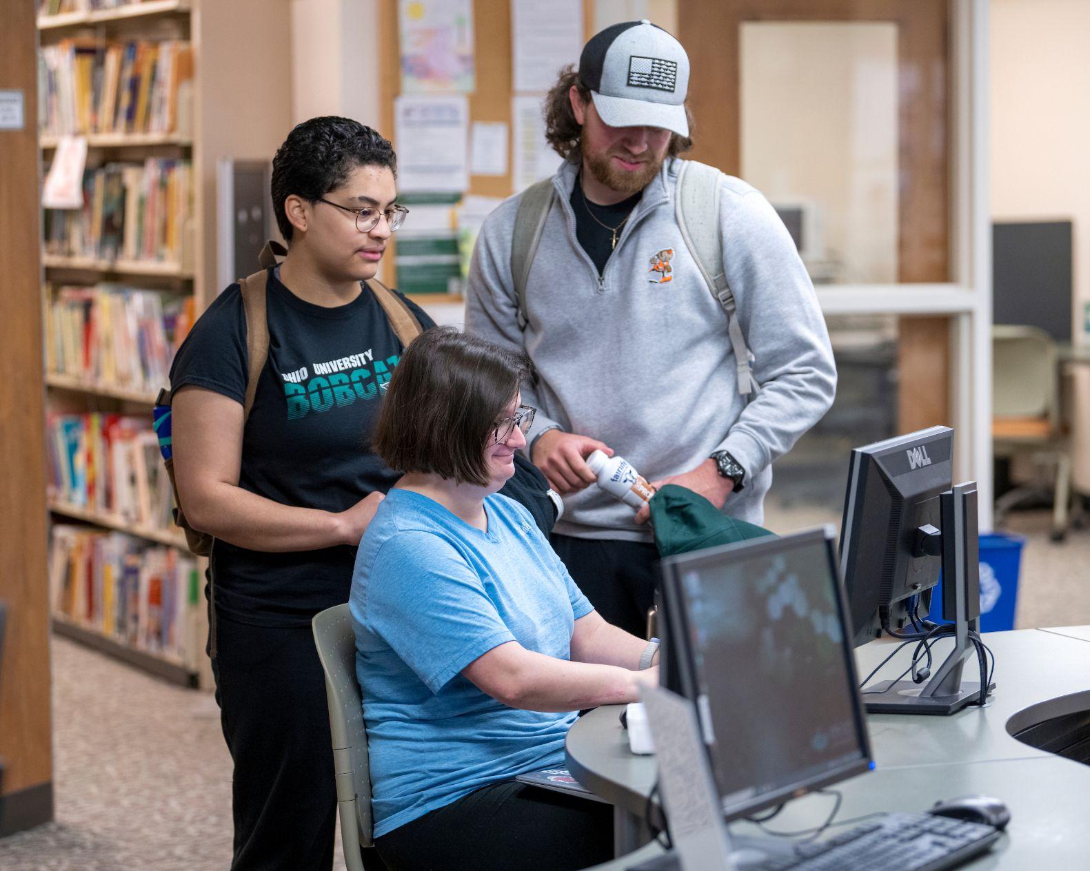 3 people, 1 male wearing a ball cap, 2 females wearing t-shirts all looking a computer screen inside the Shannon Hall library at Ohio University Eastern