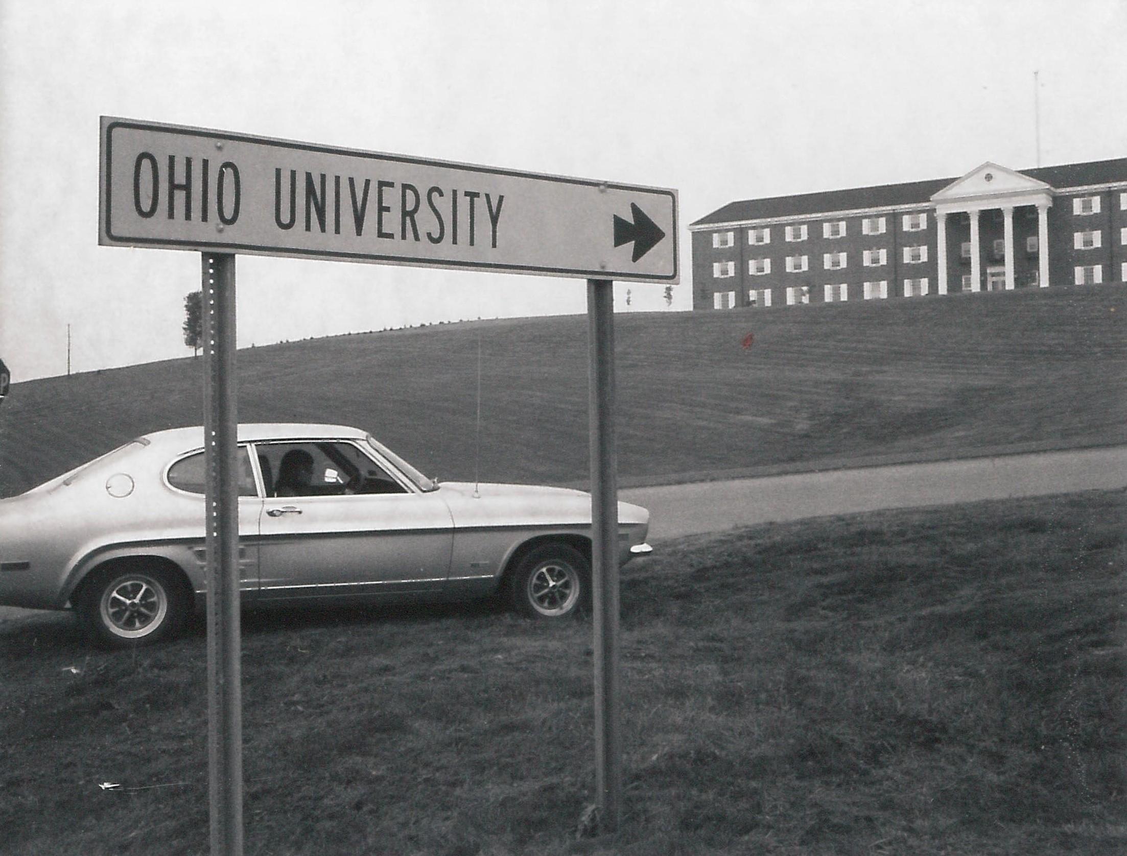 1967 vehicle on a road, next to a sign, leading up to Shannon Hall