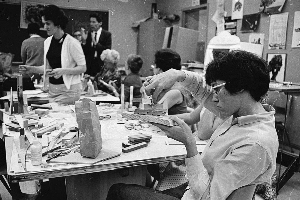Woman at a worktable manipulates wooden blocks in Ohio University Chillicothe campus art class while a man in suit gestures in the background.