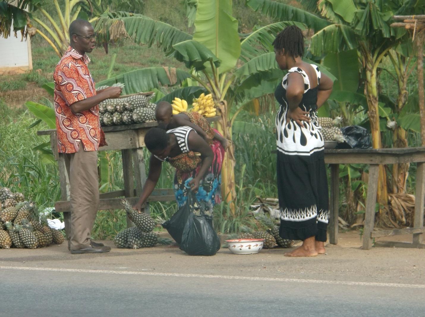 Three people standing near a market stall selling pineapples in Ghana.