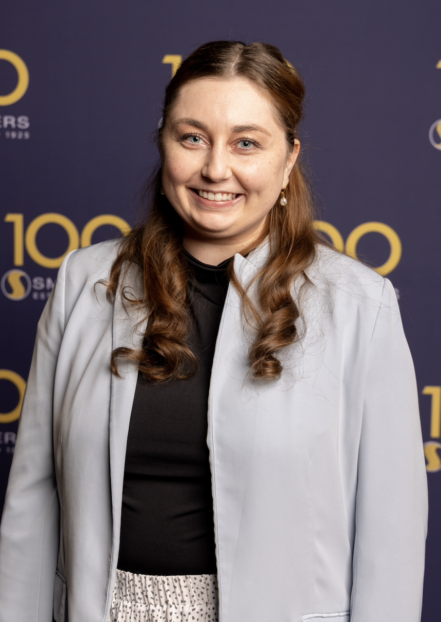 a woman facing the screen with long brown hair partially pulled back, smiling, wearing a black top under a grey suit jacket