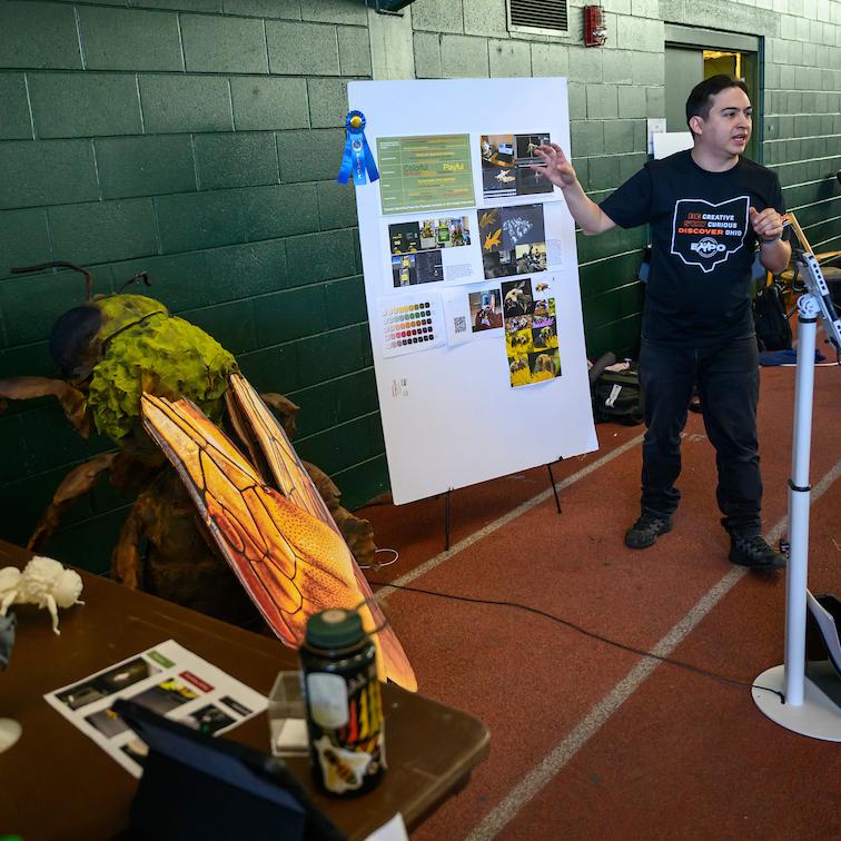 Ohio University student presenting research project at student expo involving large models and poster.