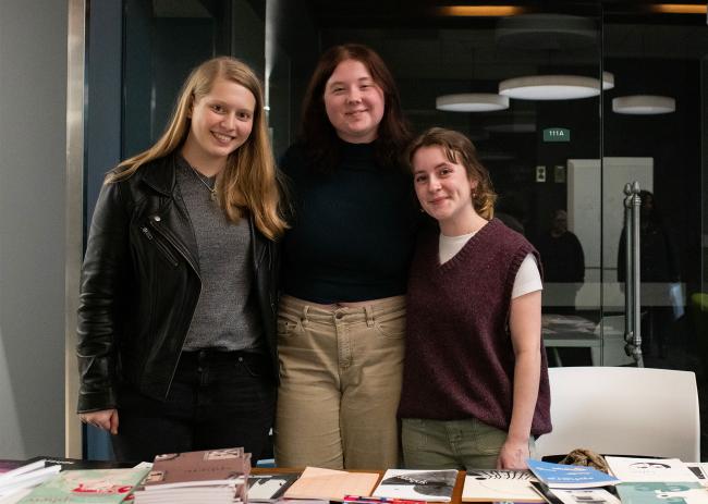 Three students pose for a photo in behind a table with books on it in Alden Library.