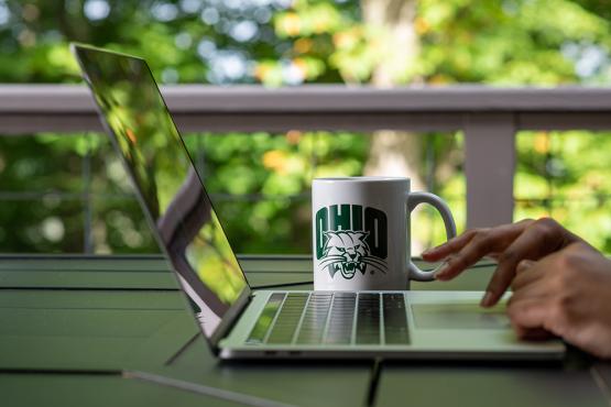 Faculty sitting outside with a laptop and a Ohio University mug.