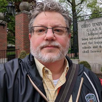 Picture of a man in yellow plaid dress shirt and Ohio University jacket standing. 