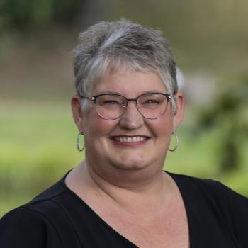 Headshot of Carleen Dotson. Woman smiling at the camera with short gray hair, dark wire framed glasses, earrings and a dark v-necked top. The background of the photo is blurred outdoor scenes with grass and trees.