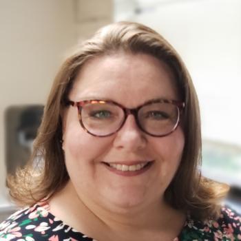 A woman smiling at the camera with chin length brown hair, brown glasses and a patterned blouse. 