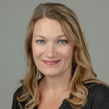 Professional Headshot of Cindi Lucas. A woman with shoulder length blonde hair smiles at the camera, she is wearing a black top and the background is a gray backdrop.